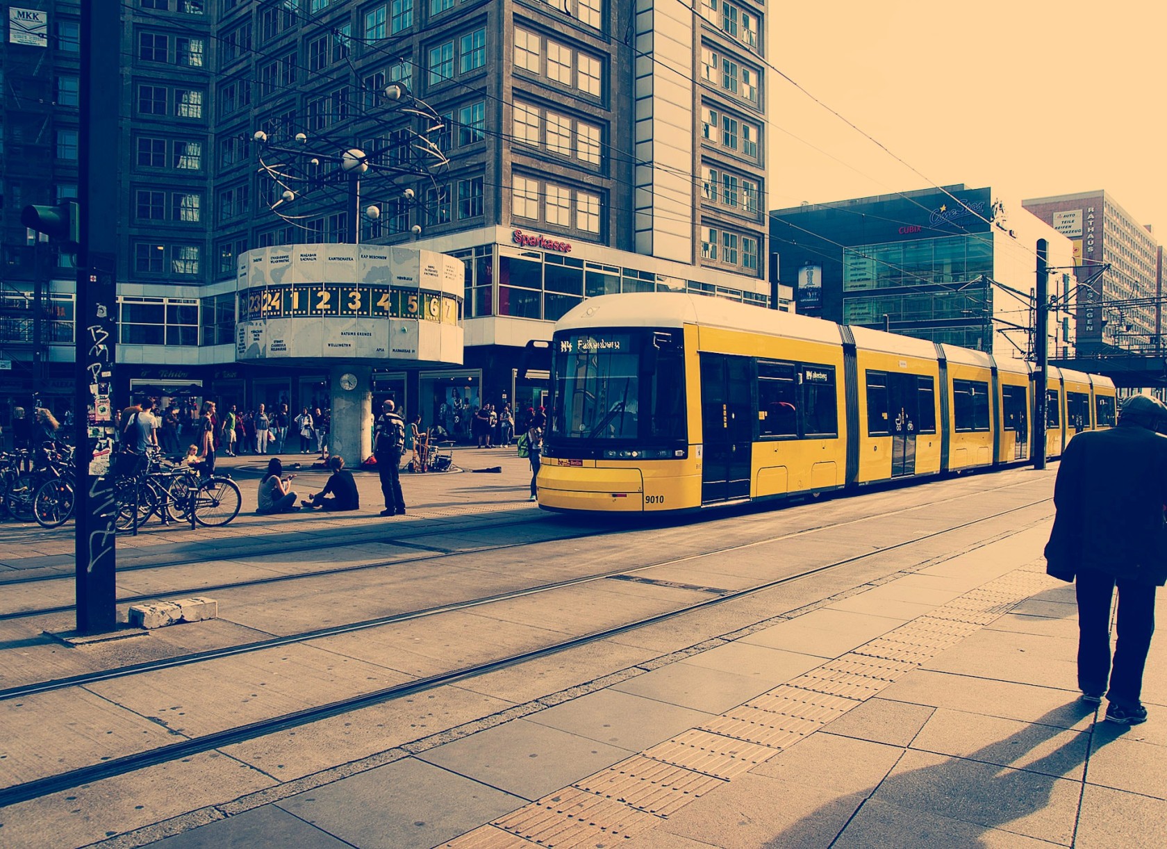 Descripción de la imagen: Paisaje urbano en la ciudad de Berlín en el que aparece un tranvía con varios edificios de fondo y algunas personas en la calle.